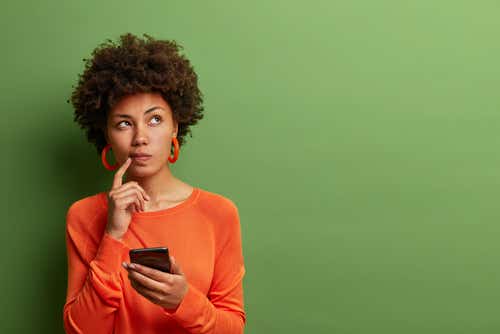 A brown-skinned woman with an Afro wears a bright orange shirt and matching earrings and stands in from of a plan green background. She has a phone in one hand and her other hand is pointing to her mouth with an inquisitive look on her face to denote confusion