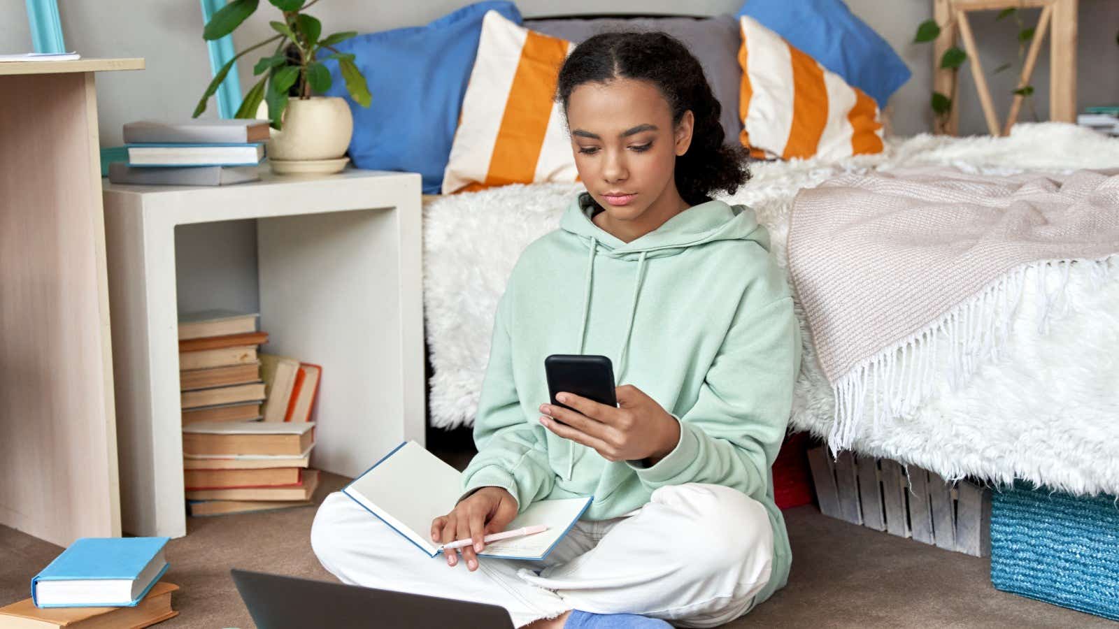 Mixed race teen school girl distant school or college student virtual remote e learning using mobile phone app and laptop in bedroom. 