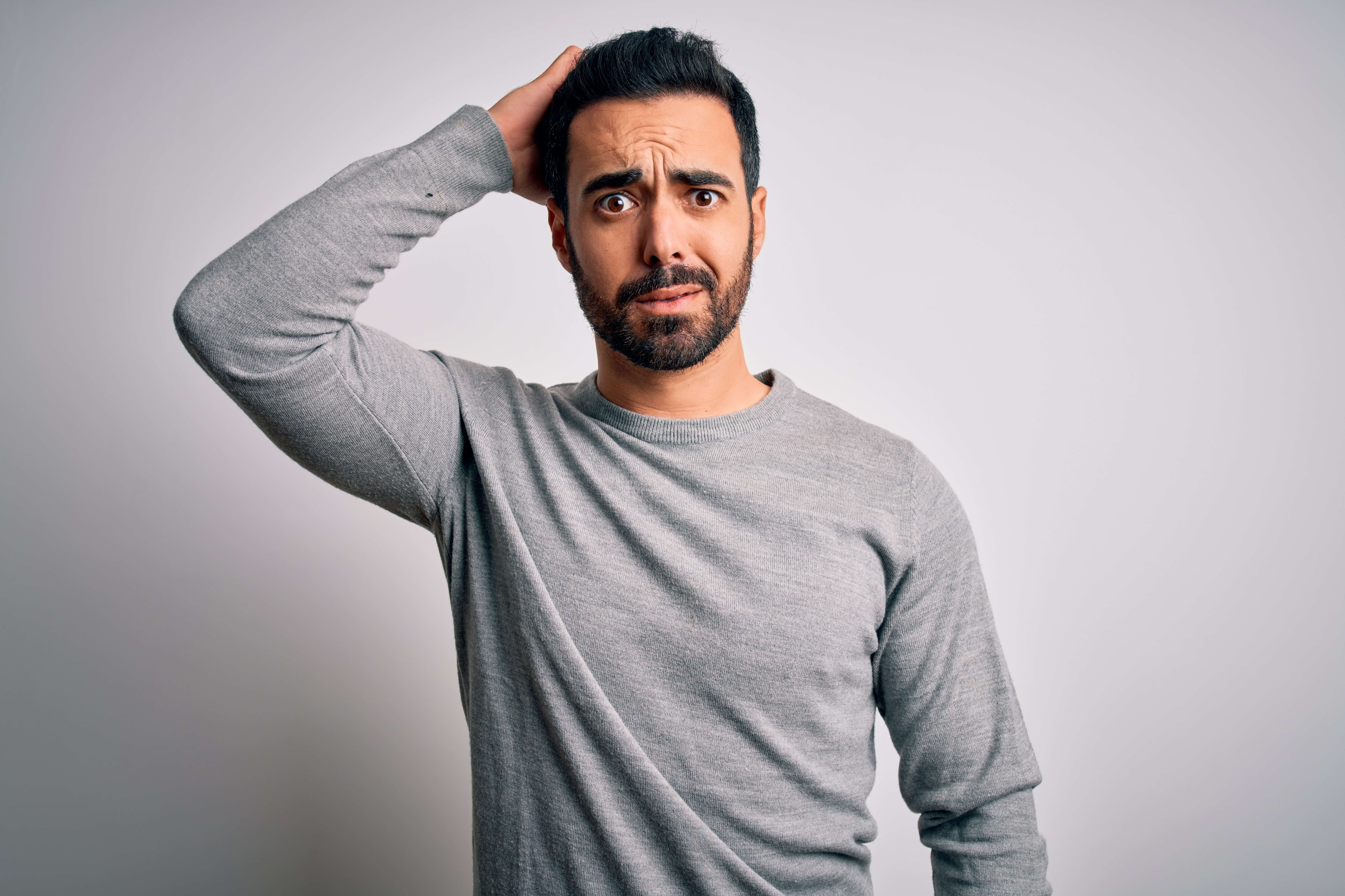 An olive-skinned man with black hair and beard and a grey sweater stands in front of a blank grey wall with his right arm raised behind his head and a look of deep confusion