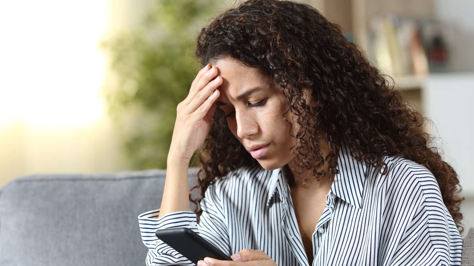 Worried woman reading on smartphone bad news sitting on a couch at home