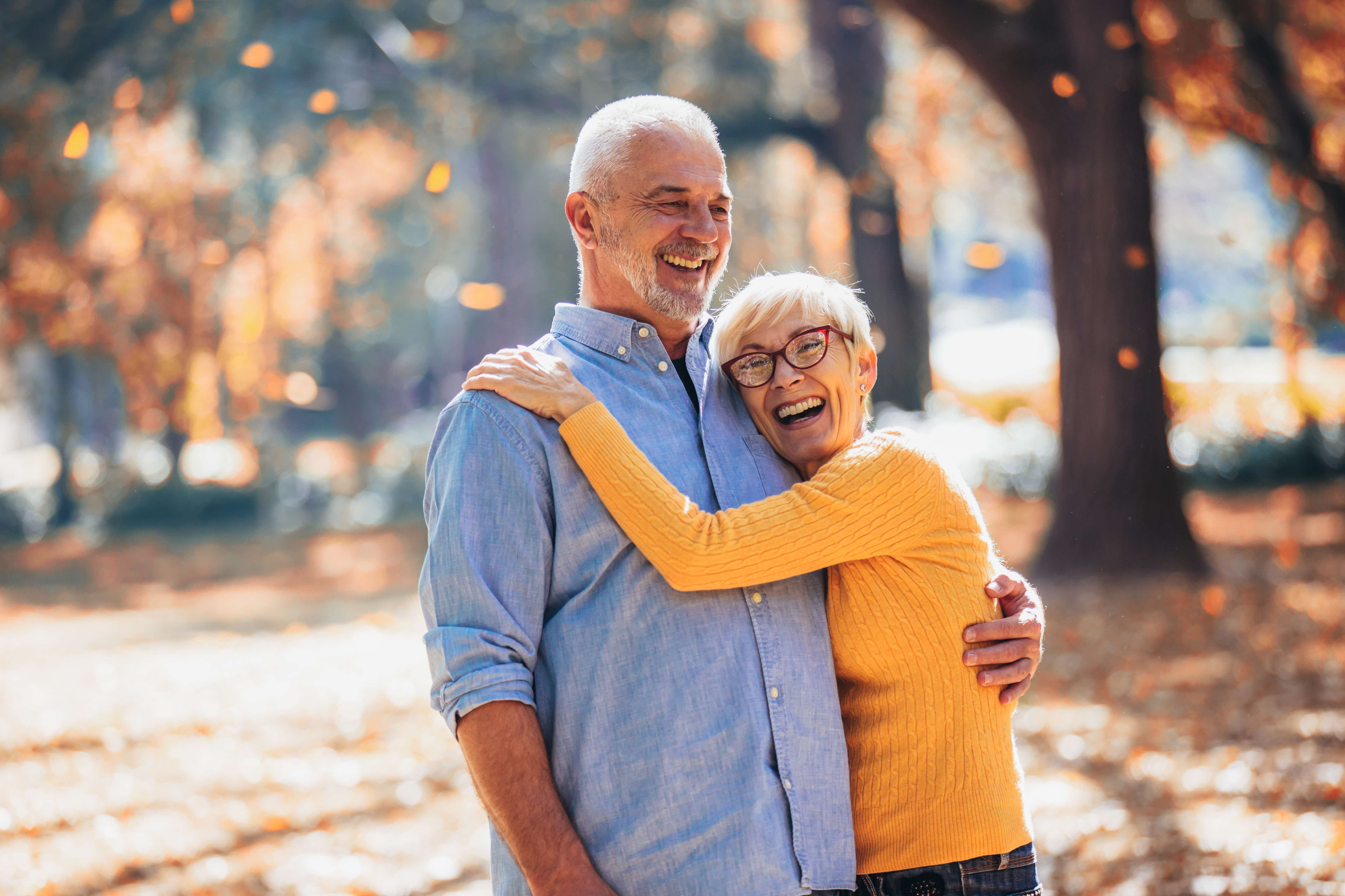 An older white male/female couple stand sideways on in front of a backdrop of autumnal trees with orange leaves falling to the ground behind them. They are laughing together