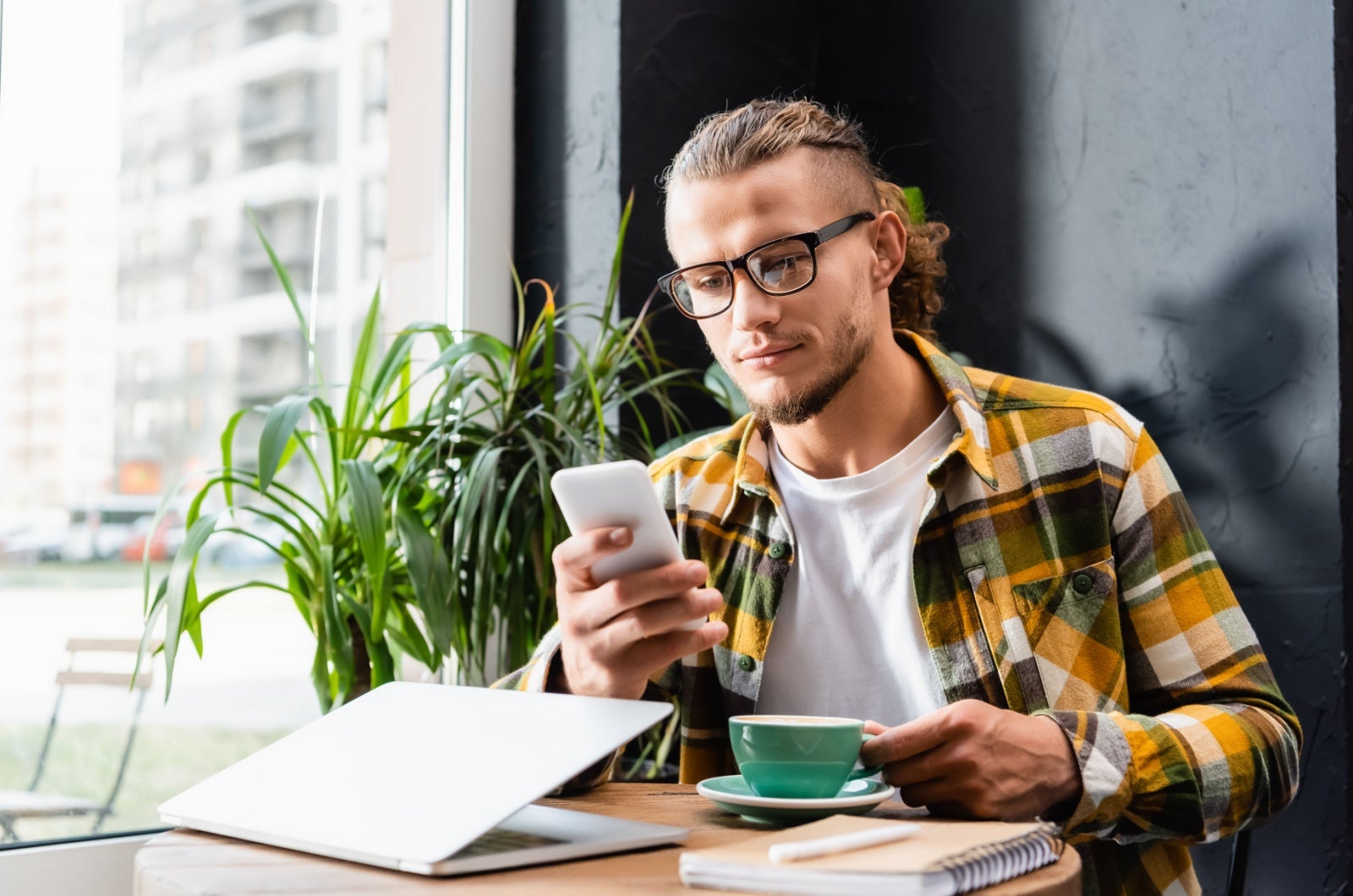 Young man in a yellow checked shirt looking at his smartphone while drinking a coffee in a cafe. In front of him is a half-closed laptop.