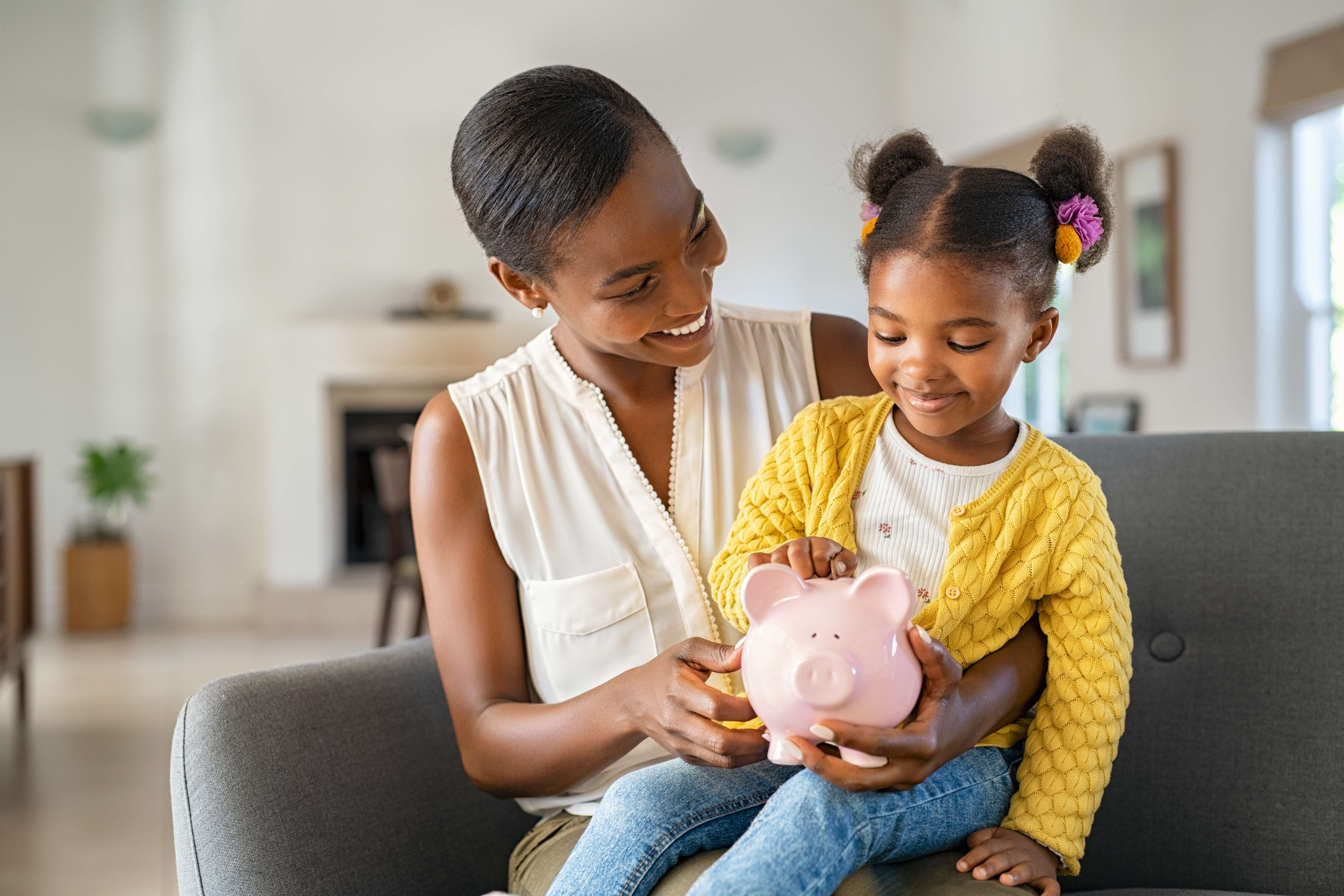 A black lady sits on a sofa in a large living room with a young girl on her lap. The girl is around 6 years old with bunches in her hair and is holding a pink pig money bank. She is putting a coin into the bank and both are smiling