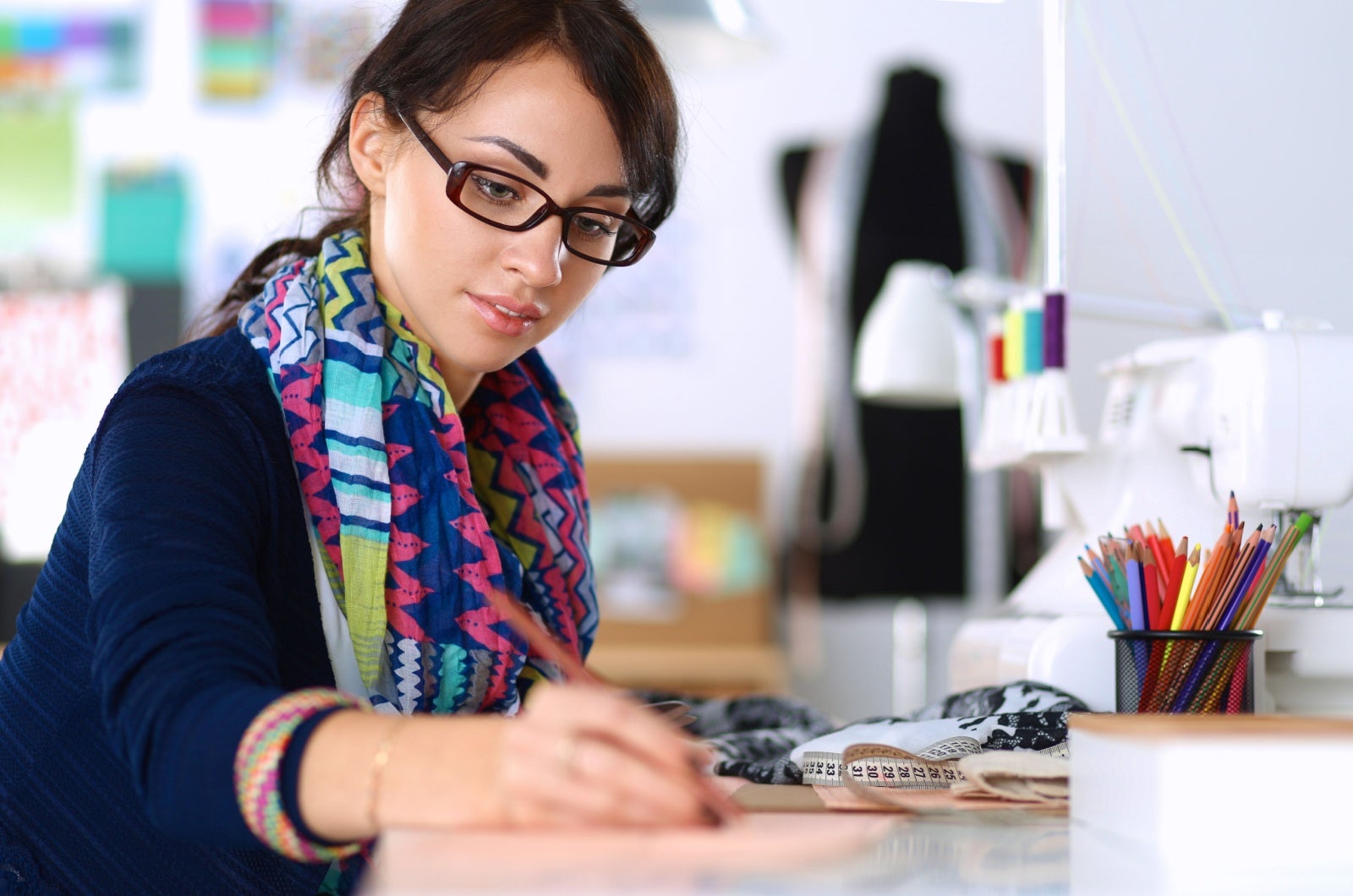 A clothes designer in a colourful scarf works at her desk with coloured pencils. In front of her is a sewing machine. A tailor's dummy is in the background. 