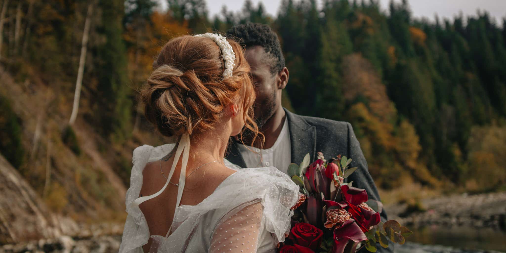An interracial couple of newlyweds walks along the shore of a mountain river. 