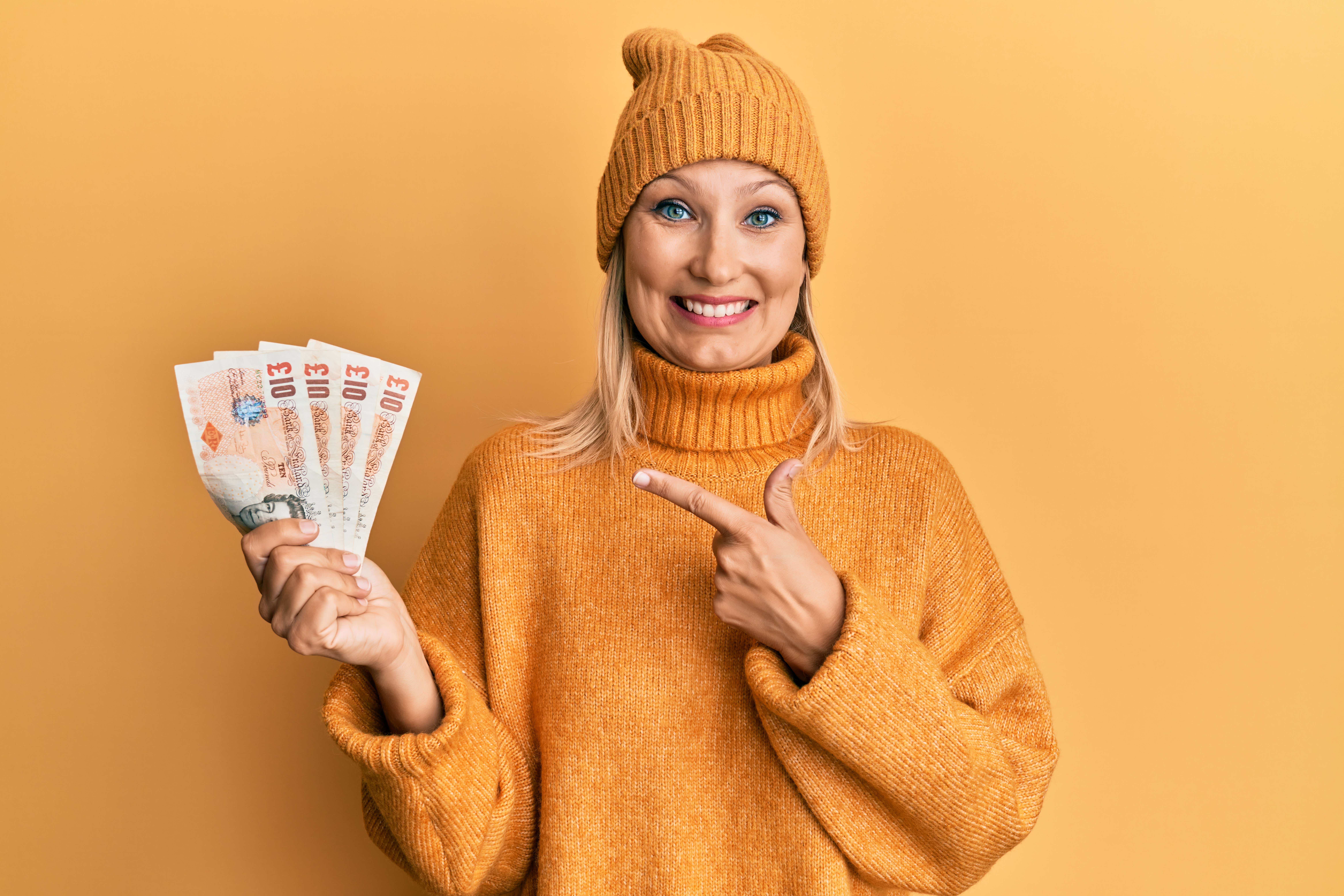 A young white woman dressed entirely in mustard yellow and standing against a mustard yellow backdrop holds a fan of UK bank notes in her right hand. He left hand is pointing to the money and she has a big smile on her face. 