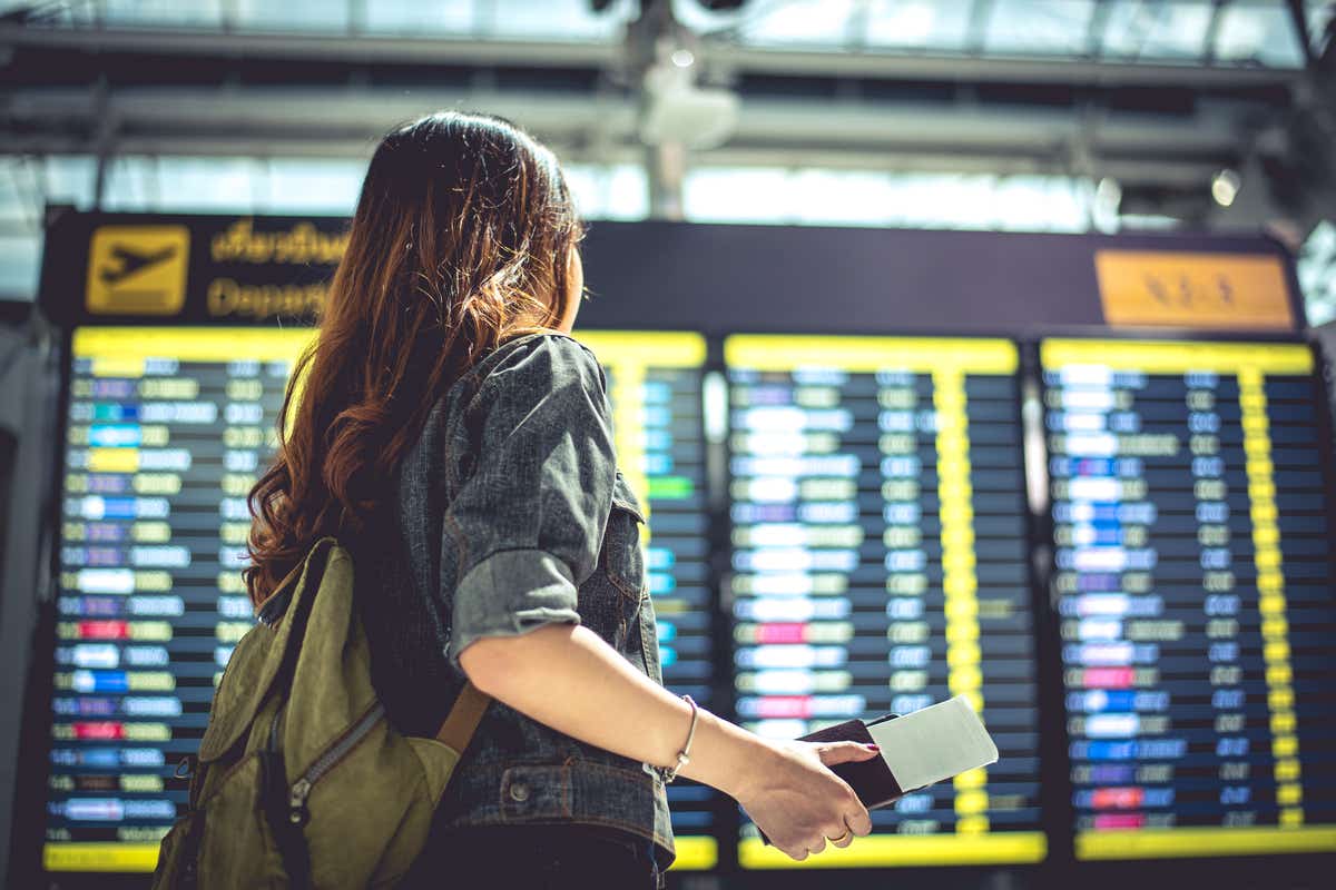 Woman carrying passport and plane tickets in front of airport departure board