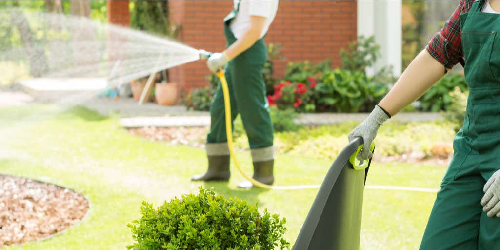 Image of two people gardening in green overalls, one person spraying house, the other person holding a chair
