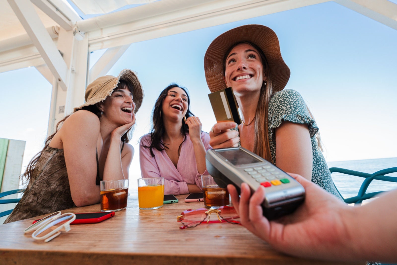 Three happy women on holiday in beachside cafe paying for drinks on credit card.
