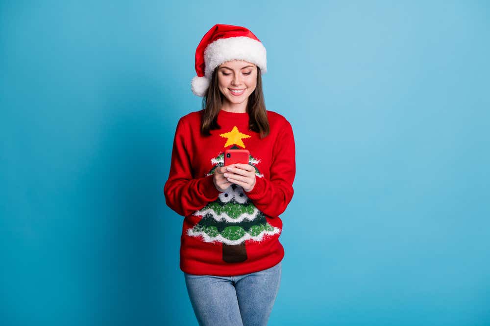 Woman is looking at her phone and is wearing a Christmas jumper and hat.
