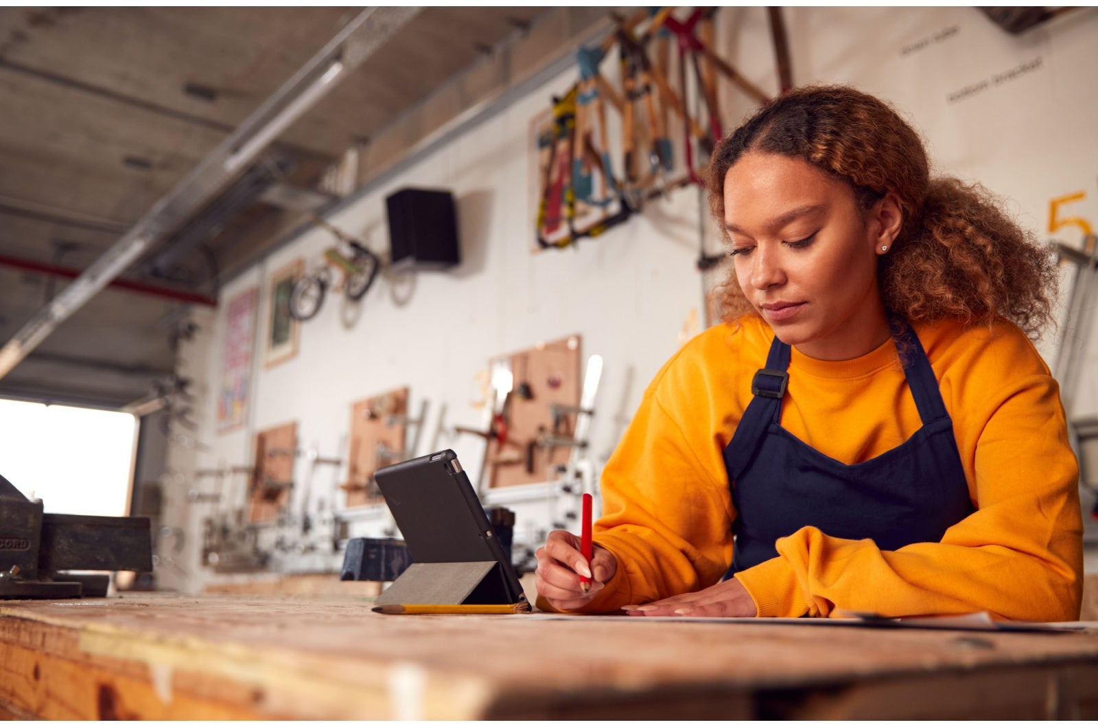 Female carpenter working with a pencil and paper. Beside her on a work bench is a tablet with keyboard. The walls are lined with her tools. 