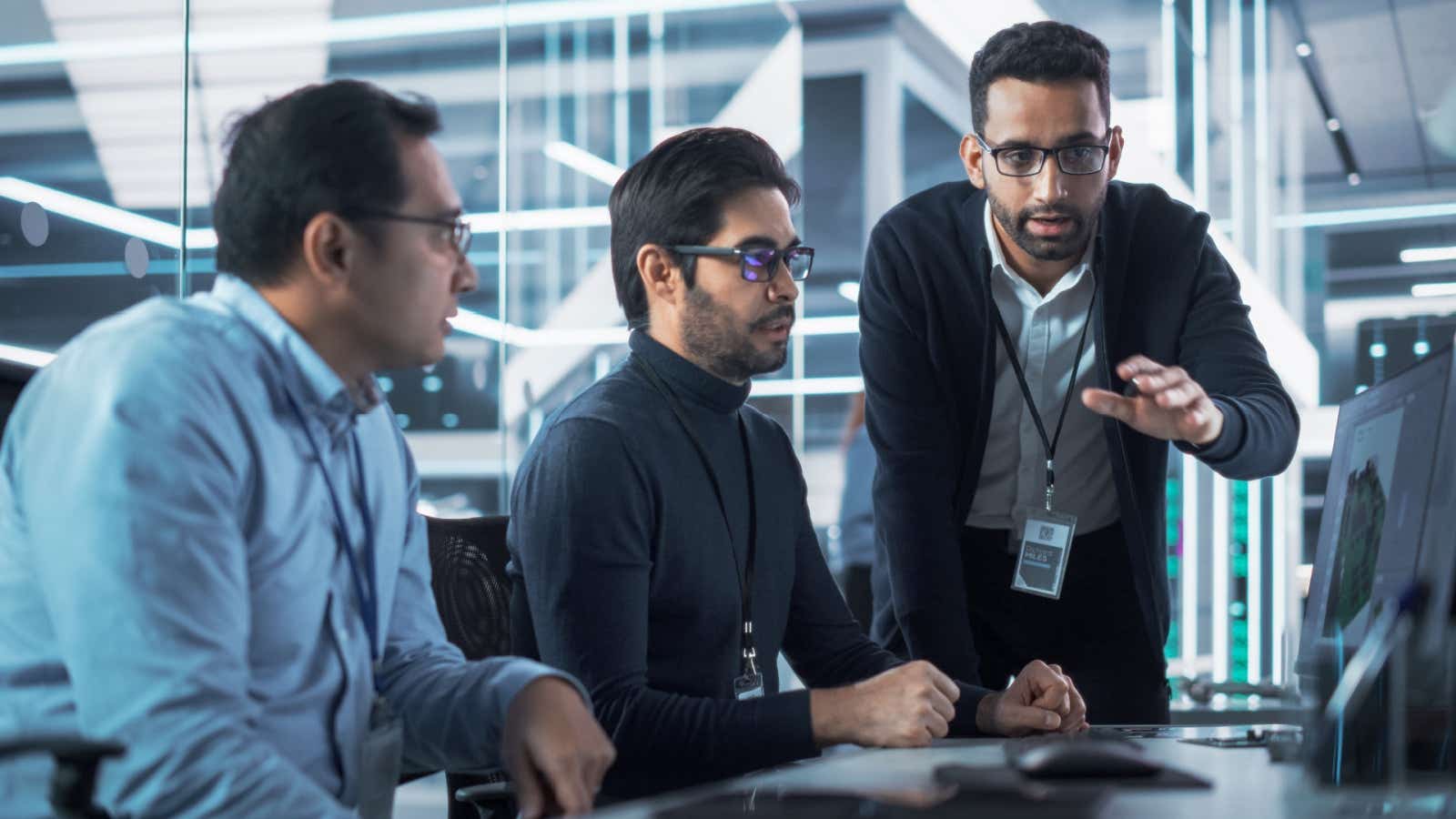 A group of workers in a modern office discussing something on one of their monitor screens