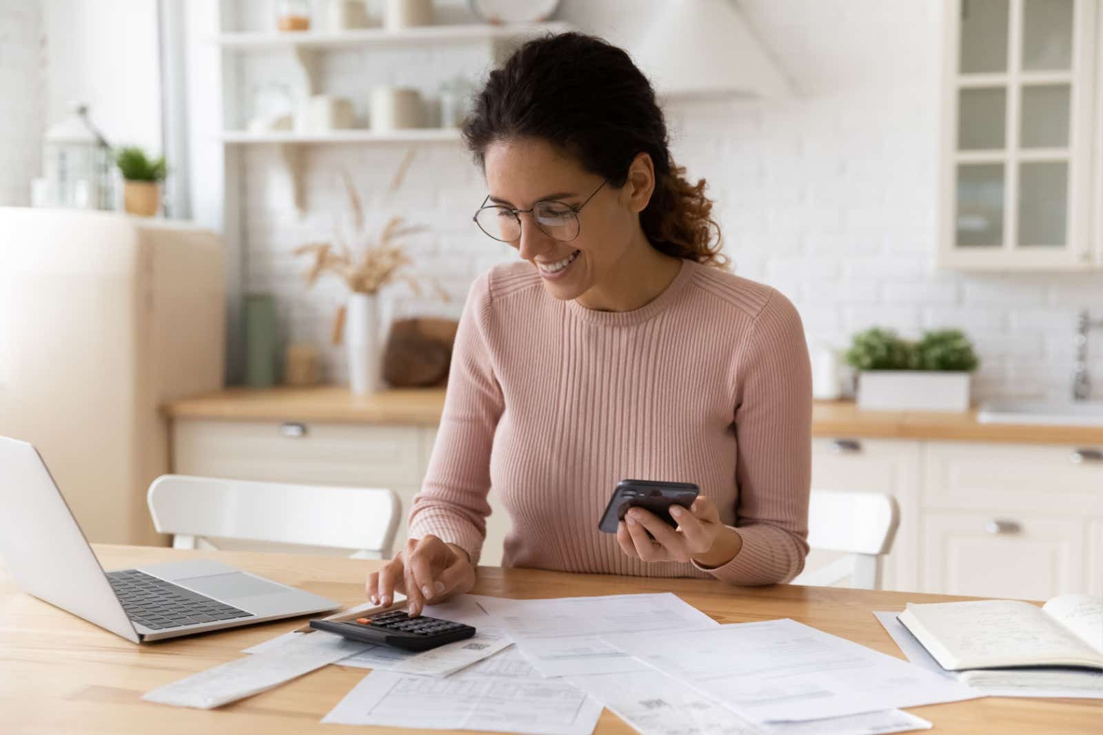 Smiling businesswoman uses desk calculator. Paperwork and a laptop lie on her desk.