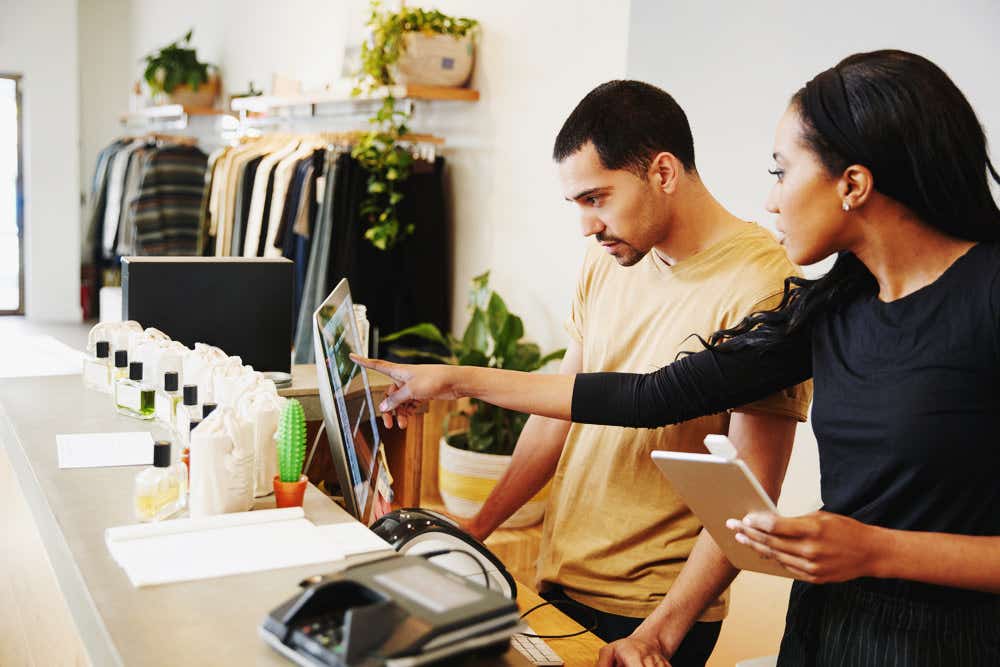 A man and a woman are in a shop behind a till looking a computer screen