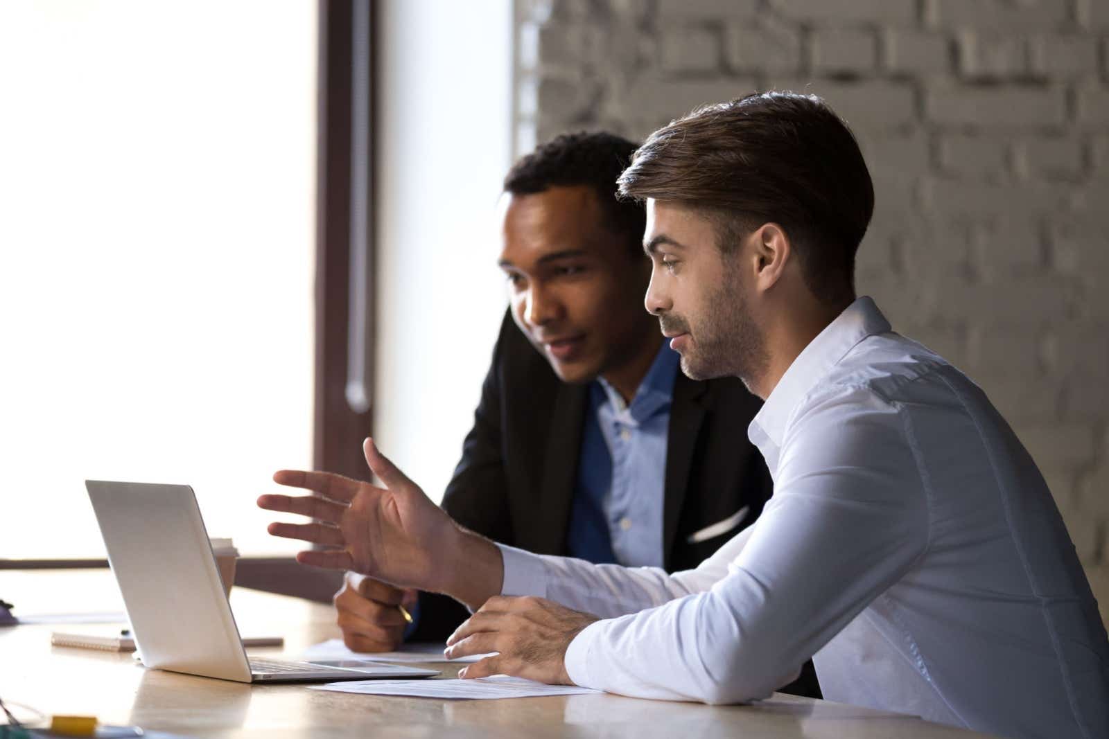 One businessman presenting ideas to another. They are seated together looking at a laptop.