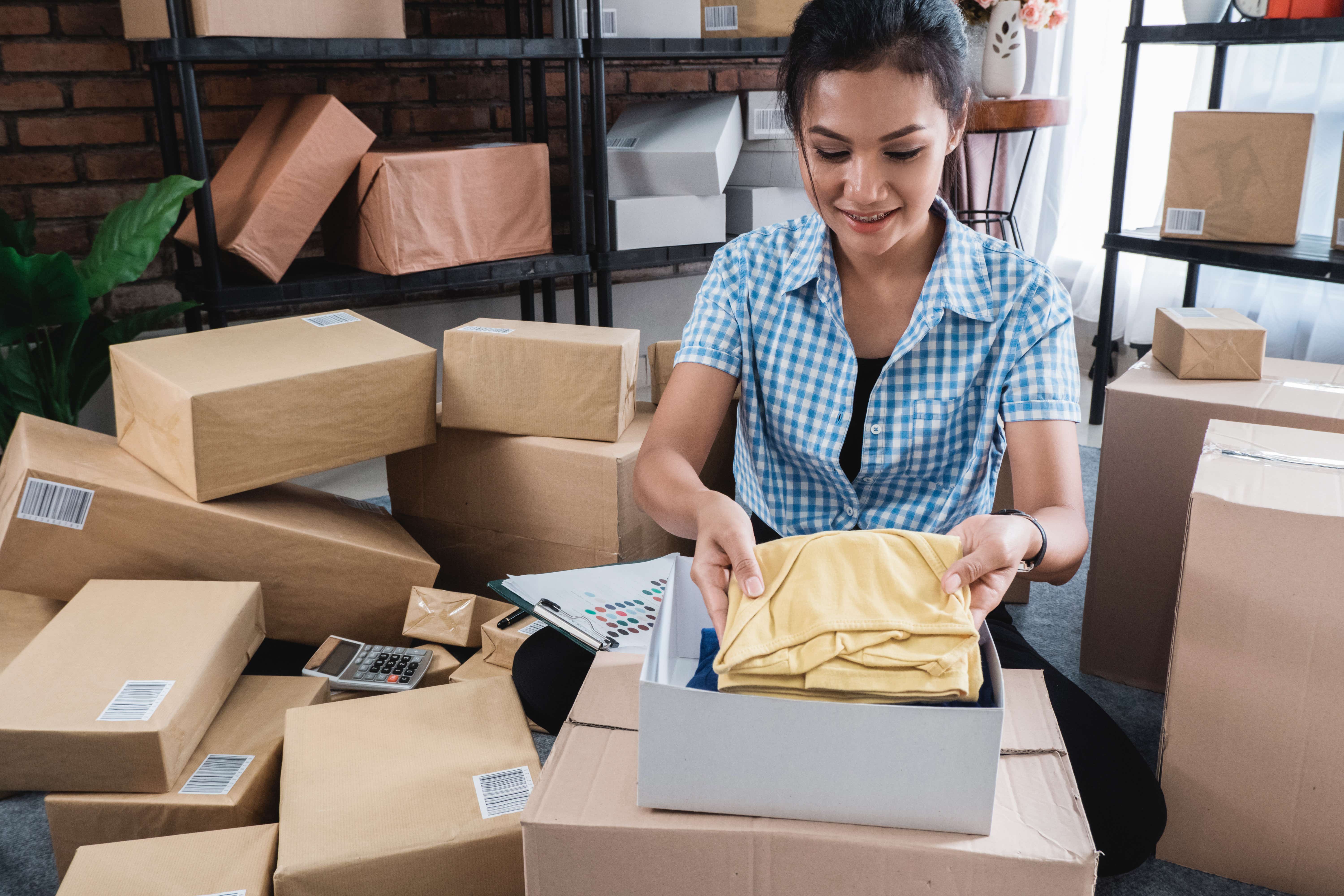A woman putting clothes into a box, surrounded by other boxes, home business, side hustle, entrepreneur, small business