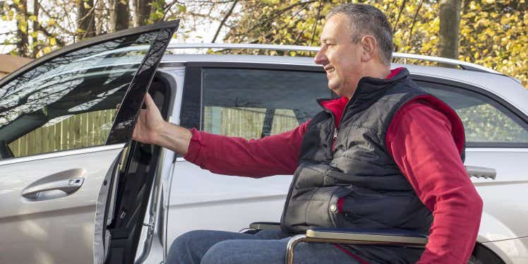 Man in a wheelchair wearing a red jumper and black gilet, opening the door of a car.
