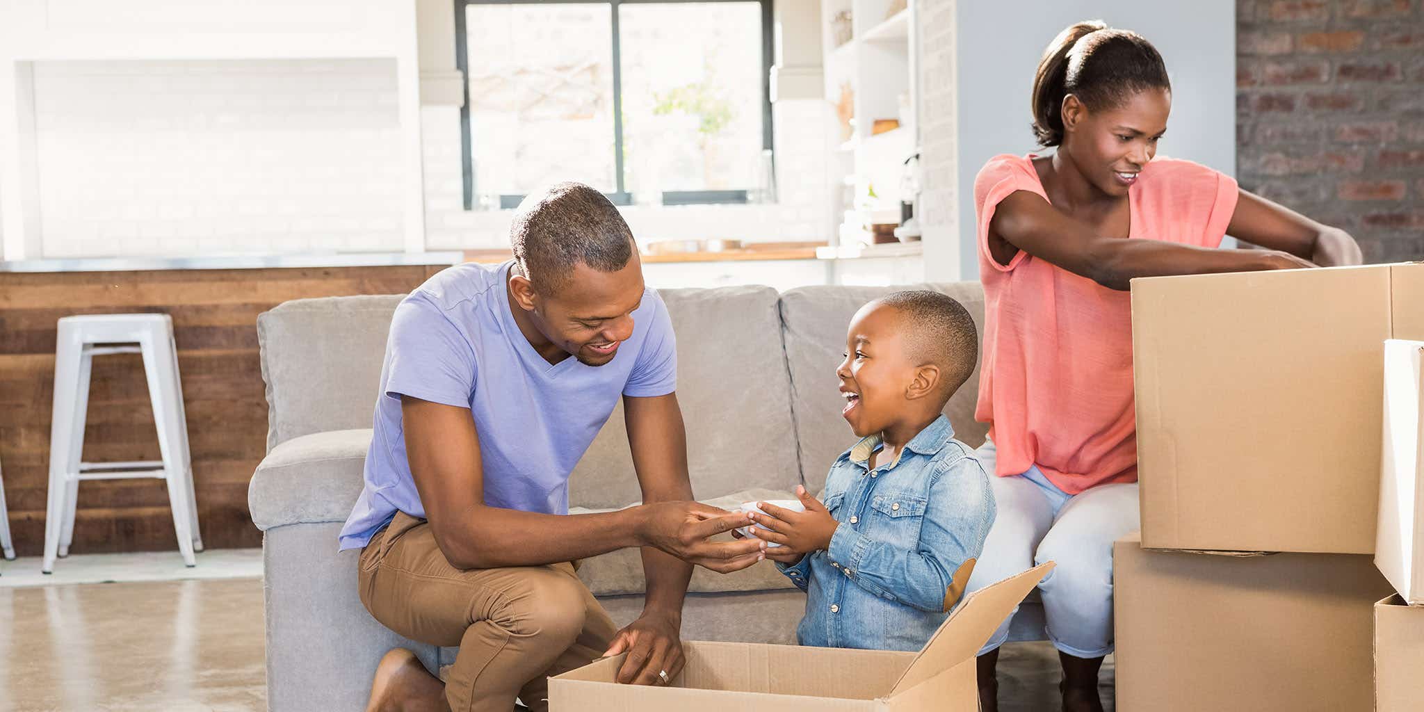 A black family of three, an adult male and female with a young boy are packing boxes to move house. All of them are smiling and there is only a sofa left in the background, with multiple boxes in the foreground of the room