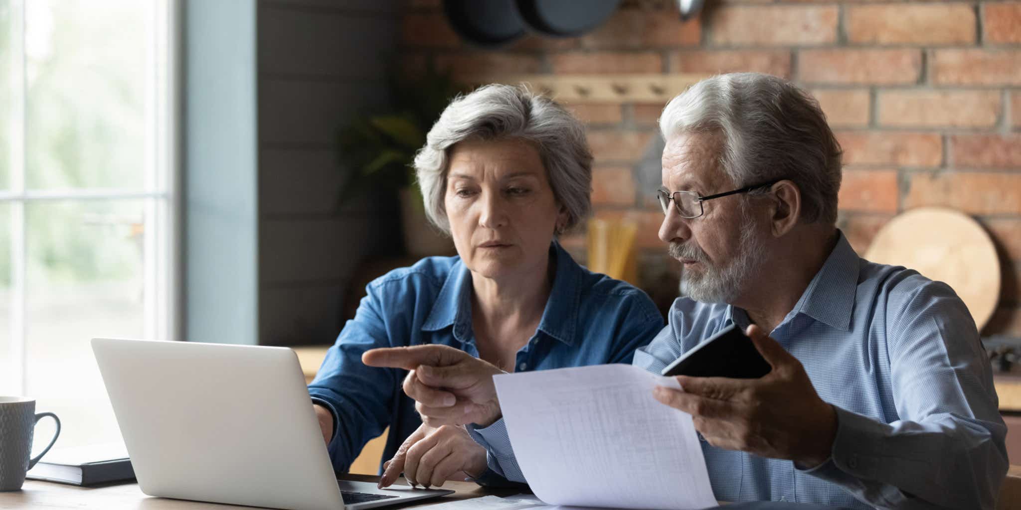 Senior husband and wife sit at table at home look at laptop screen