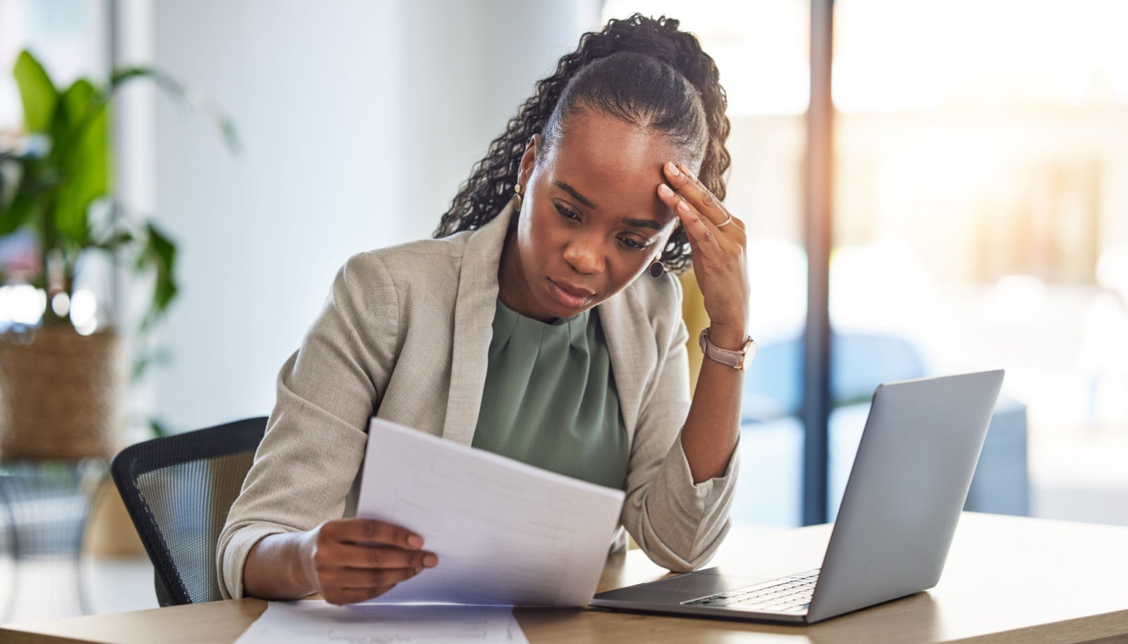 A worried looking businesswoman sits at her desk, looking at a letter she's holding. 