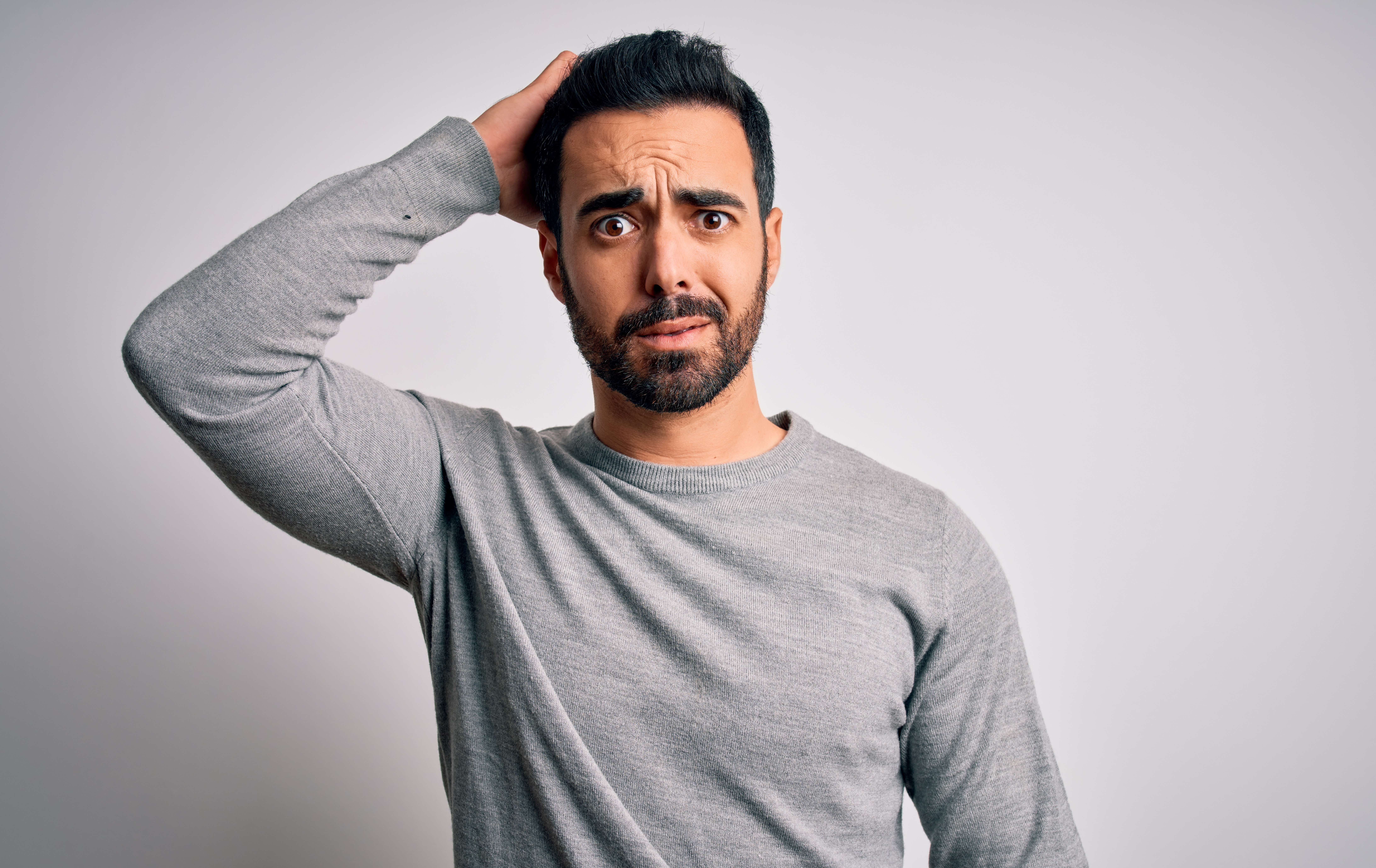 An olive-skinned man with black hair and beard and a grey sweater stands in front of a blank grey wall with his right arm raised behind his head and a look of deep confusion