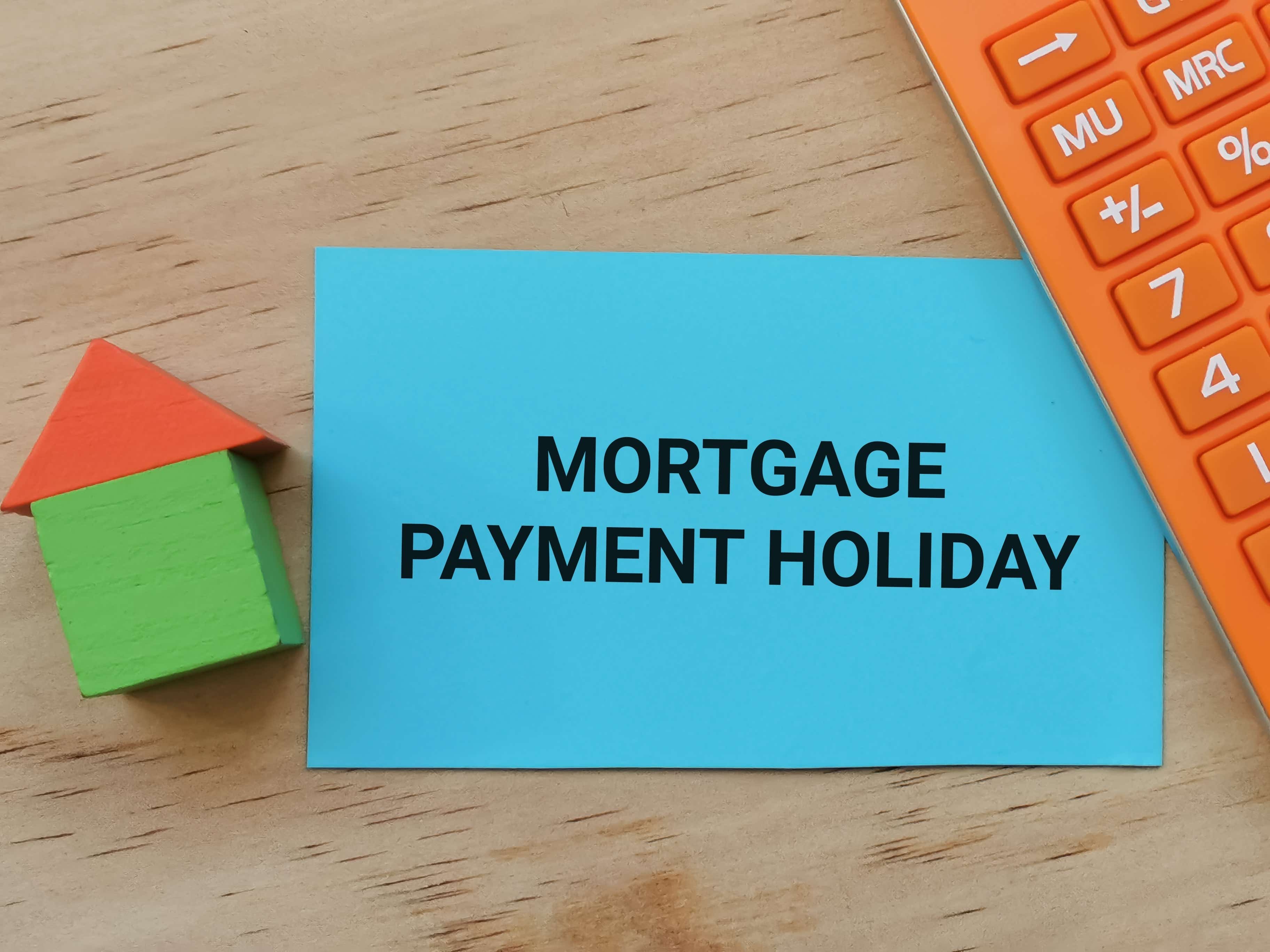On a wooden desk lays an orange calculator, a small model of a green house with a red roof and a blue card with the words 'mortgage payment holiday' in bold print.