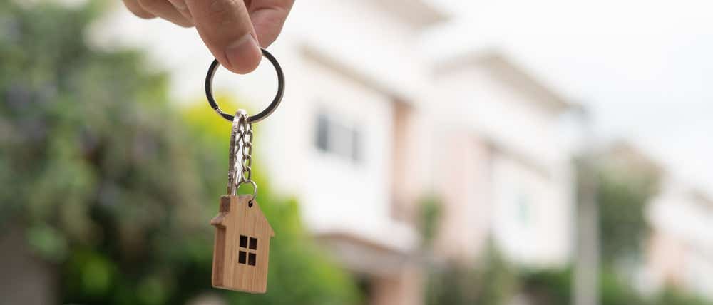 Picture of a blurred house with a someone holding a keychain with a wooden house.